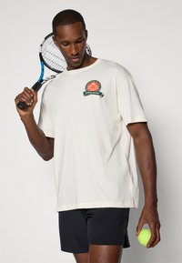 Kremowa bawełniana koszulka z okrągłym dekoltem i haftowanym logo. W jednej ręce trzymane są czarne spodenki i rakieta tenisowa, a w drugiej piłka tenisowa.