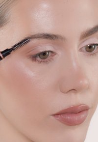 Brosse de mascara appliquant le produit sur des sourcils naturels, légèrement fournis ; le modèle a une peau lisse avec un illuminateur subtil et du rouge à lèvres nude.
