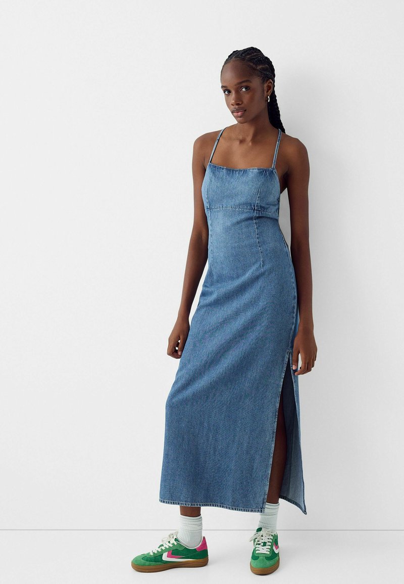 Abito maxi in denim con spalline sottili, caratterizzato da una silhouette dritta, uno spacco laterale e una texture di tessuto azzurro chiaro. Accentuato da sneakers sportive verdi.