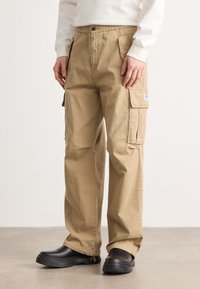 Khaki cargo pants with an elastic waistband, side pockets, and a textured fabric. Worn with black clogs, standing on a neutral surface.