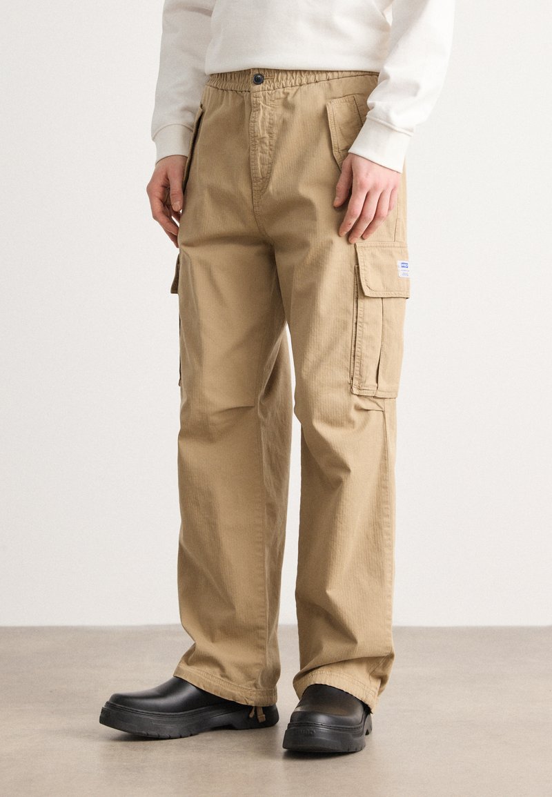 Khaki cargo pants with an elastic waistband, side pockets, and a textured fabric. Worn with black clogs, standing on a neutral surface.