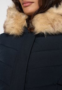 Giacca imbottita nera con collo in pelliccia marrone chiaro morbida, caratterizzata da una texture liscia e un design trapuntato diagonale sul petto.