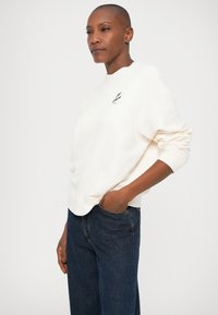 Felpa crema realizzata in morbido tessuto, con collo rotondo, polsini a costine e un piccolo dettaglio del logo nero. Indossata con jeans blu scuro.