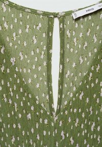 Gros plan d'une robe en tissu vert à motifs floraux, avec de petites fleurs blanches et lavande, une fente en V dans le dos et une étiquette visible en haut.
