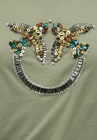 Δύο συμμετρικές πεταλούδες με πολύτιμους λίθους κεχριμπαριού, πράσινους και χρυσούς λίθους και παγιέτες σε πράσινο ύφασμα.