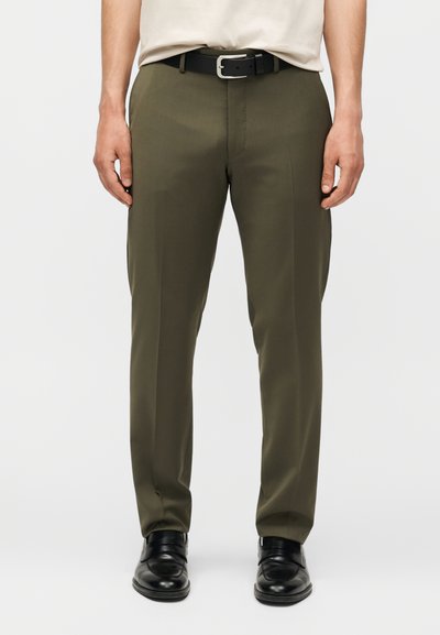 Homme portant un pantalon vert olive sur mesure, une ceinture noire, des chaussures habillées en cuir noir et une chemise beige rentrée, debout devant un fond uni.