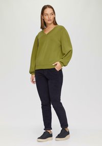 Olijfgroene, v-hals pullover met lange, ballonmouwen; gecombineerd met donkere broek en zwarte sneakers met een gestructureerde zool.