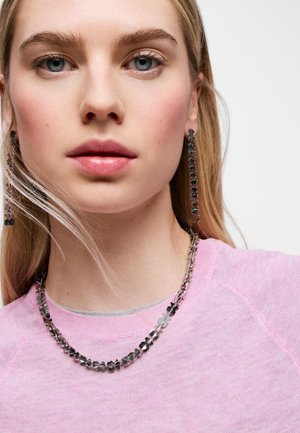 Giovane donna con lunghi capelli biondi indossa una collana in argento con motivi floreali e lunghi orecchini abbinati, vestita con un top rosa chiaro.