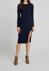 Laivastonsininen pitkähihoinen mekko sivuhalkiolla, valmistettu sileästä kankaasta. Yhdistetty ruskeisiin käärmeennahkakuosisin nilkkureihin.