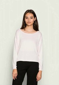 Blusa de manga longa rosa clara com um decote largo, corte descontraído e textura suave, combinada com calças pretas. Design simples e minimalista.