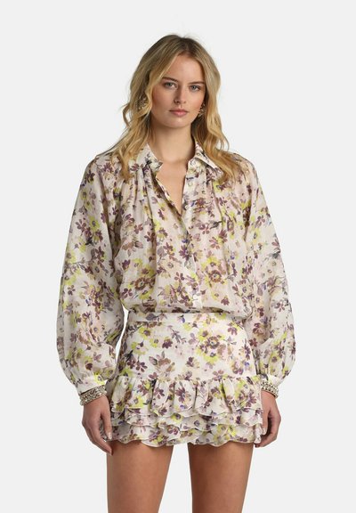 Kvinde iført en langærmet blomsterbluse med knapper og matchende flæset mininederdel, stående mod en ensfarvet baggrund.