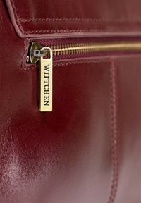 Burgundowa skórzana torebka z bliska z złotym zamkiem błyskawicznym i metalową zawieszką z wygrawerowanym napisem "WITTCHEN". Gładka tekstura z drobnymi detalami ściegów.