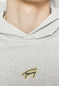 Un sweatshirt gris en tissu doux, doté d'une capuche avec cordon de serrage et d'un logo brodé en noir et or sur la poitrine.