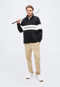 Νέος άνδρας που κρατάει μια ράβδο γκολφ στον ώμο, φοράει μαύρο-λευκό φούτερ Jordan, μπεζ παντελόνι και λευκά αθλητικά παπούτσια, στέκεται σε απλό φόντο.