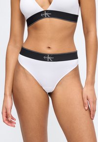 Γυναίκα που φοράει λευκό bralette Calvin Klein και σλιπ με ψηλή μέση με μαύρες ελαστικές ταινίες και λογότυπο σε μονόχρωμο φόντο.