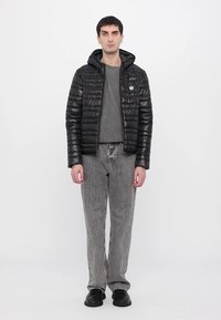 Chaqueta de plumas negra con capucha y diseño acolchado, sobre una camiseta gris. Jeans grises y zapatos negros completan el atuendo.