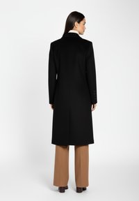 Sort lang frakke med en struktureret design, spidse lapeller og en midtervent, båret over beige vide bukser. Glat stoftekstur.