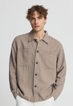 Giovane uomo con capelli ricci che indossa una camicia taupe con bottoni e tasche sul petto, pantaloni bianchi, in piedi con le mani nelle tasche.