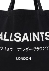 Black cotton tote bag featuring large white text "ALLSAINTS" and additional lettering in Japanese, with "LONDON" at the bottom.