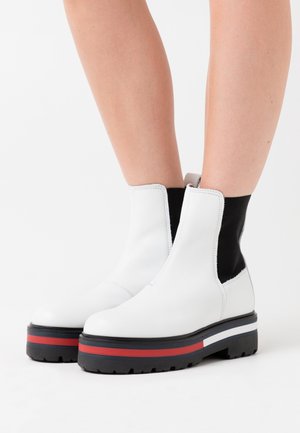 Bottines blanches en cuir avec des côtés élastiques noirs, semelle épaisse noire accentuée de rayures rouges et blanches. Texture lisse et design à bout arrondi.