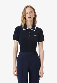 Polo shirt en ribbed marine avec col et poignets blancs, comportant trois boutons et le logo Lacoste. Stylé avec un pantalon taille haute marine.