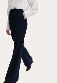 Marineblauwe streepjesbroek met een hoge taille, rechte pijpen en zijzakken, gecombineerd met een witte blouse met lange mouwen en bordeauxrode schoenen.