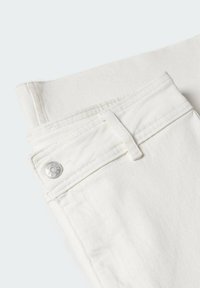 Gros plan sur un jean en denim blanc cassé plié, montrant la ceinture, la boucle de ceinture et un bouton argenté avec un texte gravé.