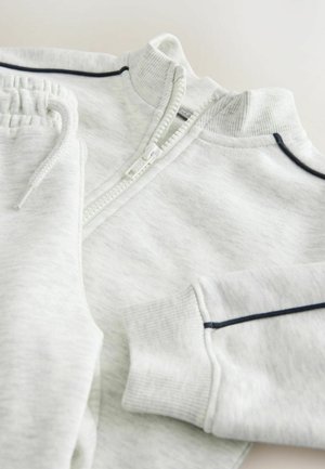 Lichtgrijze trui met een hoge kraag, geribbelde manchetten en ritssluiting. Heeft marineblauwe accenten langs de schoudernaden en op het zakgebied.