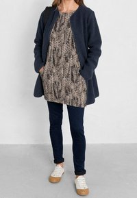 Marinblå fleecejacka, mönstrad beige klänning med bladdesign, mörkblå jeans, vita sneakers med bruna detaljer, ställande pose.