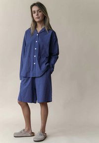 Navyblauwe katoenen pyjama's met witte stippen, bestaande uit een overhemd met knopen en knielange shorts, gecombineerd met grijze instapklompen.
