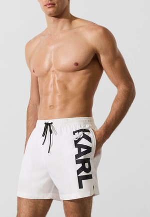 Homme torse nu portant un short de bain blanc avec un cordon noir et un grand texte noir « KARL » sur la jambe gauche, main dans la poche.