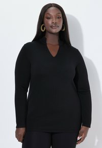 Zwarte lange mouwen trui met een V-hals en ribbeltextuur. Het materiaal lijkt glad, gecombineerd met zwarte leggings en grote ronde oorbellen.