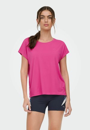 Kvinde med langt mørkt hår bundet tilbage bærer en løs pink kortærmet skjorte og sorte sportsshorts med en hvid sideliste.