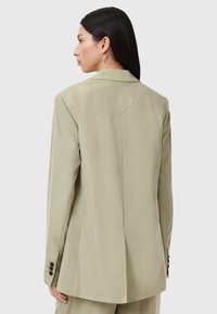 Blazer su misura verde chiaro realizzato in tessuto morbido. Presenta una sola apertura sul retro e tre bottoni neri su ciascuna manica.