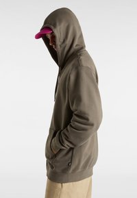 Olivgrüner Hoodie mit Kanguru-Tasche und taillierter Passform. Akzentuiert durch eine rosa Kappe, die verkehrt herum getragen wird.