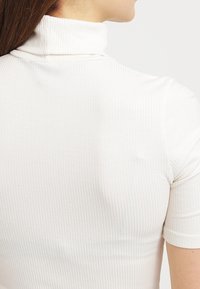 Maglia bianca a collo alto a costine con maniche corte, caratterizzata da una vestibilità aderente e un tessuto testurizzato. Il design pulito enfatizza la semplicità e la versatilità.
