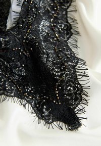 Melna mežģīņu apmale ar ziedu rakstiem, pērlītēm un nograuztām malām, novietota uz gludas baltas auduma.