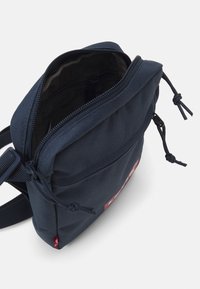 Borsa a spalla in tessuto blu navy con zip superiore, tracolla regolabile e un'etichetta con logo rosso. Presenta una superficie esterna testurizzata e dettagli con cordoncino.