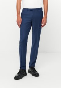 Ein Mann trägt marineblaue Anzughose, schwarze Schnürschuhe und ein weißes Hemd und steht vor einem schlichten weißen Hintergrund.