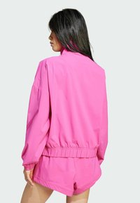 Chaqueta de tiro alto de color rosa con cuello alto, cintura elástica y corte holgado. Tejido ligero con textura suave y mangas largas.