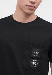 Άνδρας που φοράει μαύρο t-shirt με τσέπη στο στήθος με επιθέματα Armani Exchange που αναγράφουν A|X Armani Exchange και Milano 1991 New York.