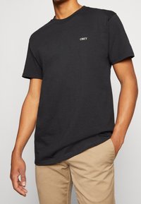 Svart bomulls-T-shirt med korta ärmar och rund hals, med en liten vit "OBEY"-logotyp på bröstet.