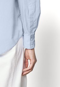 Person som bär en ljusblå långärmad skjorta med knäppt manschett och vita byxor, med handen avslappnad vid sin sida mot en vit bakgrund.
