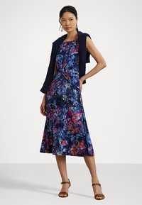 Abito a fiori in navy con dettagli in rosa, rosso e verde. Silhouette a forma di A, tessuto morbido e dettaglio alla vita annodato. Abbinato a un gilet navy.
