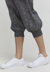 Svarta mönstrade joggers med elastiska avslut, som är kombinerade med vita sneakers med en gul accent på tungan. Detaljerad blommönster synlig.