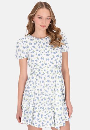Fiatal nő hullámos hajjal, fehér, rövid ujjú ruhában, kék bogyómintákkal és megkötött derékövevel, semleges háttér előtt állva.