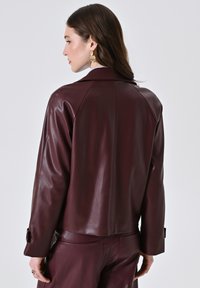 Vrouw met lang bruin haar, draagend gouden oorbellen en een donkere bordeaux leren jas met bijpassende broek, die zijwaarts staat tegen een effen achtergrond.