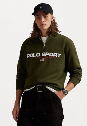 Olive green zip-up sweatshirt with "POLO SPORT" logo in white and red. Worn over a white shirt, paired with black pants and a watch.