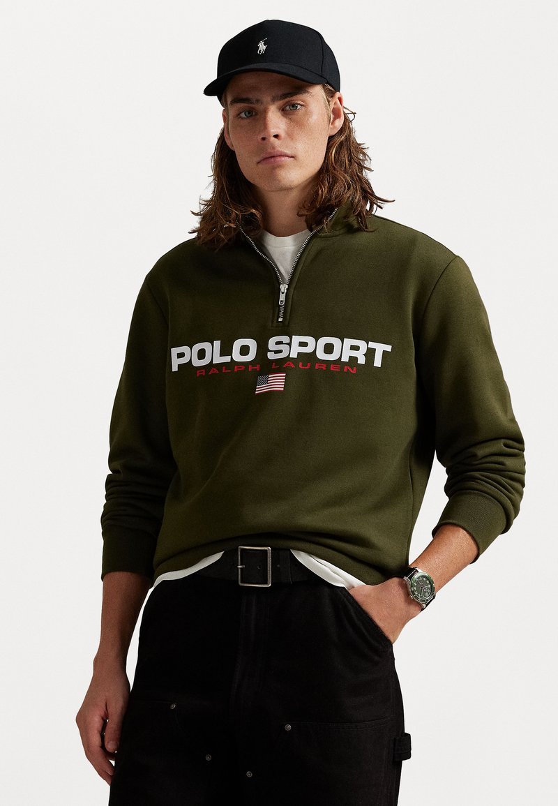 Olive rohelise värviga lukuga fliis, millel on valge ja punane "POLO SPORT" logo. Kandev valge särk, mustade pükste ja kellaga.