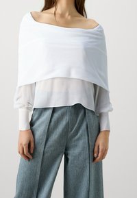 Haut blanc à épaules dénudées superposé à une blouse blanche transparente, associé à un pantalon en denim bleu à taille haute et à jambe large avec une texture subtile.